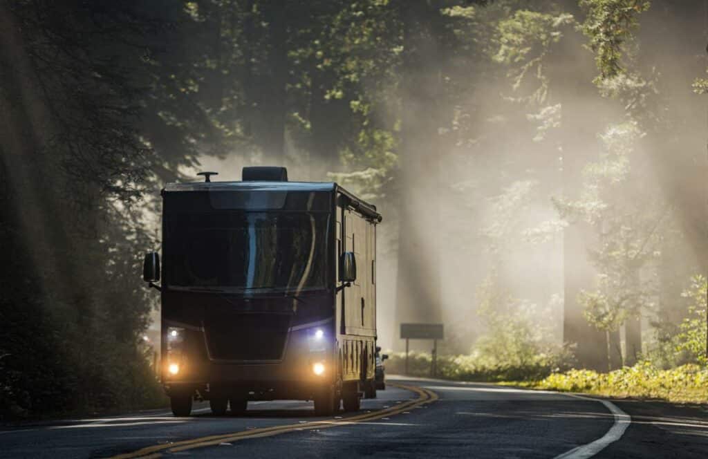 An RV drives around a curve with its headlights on in a lush forest with morning fog