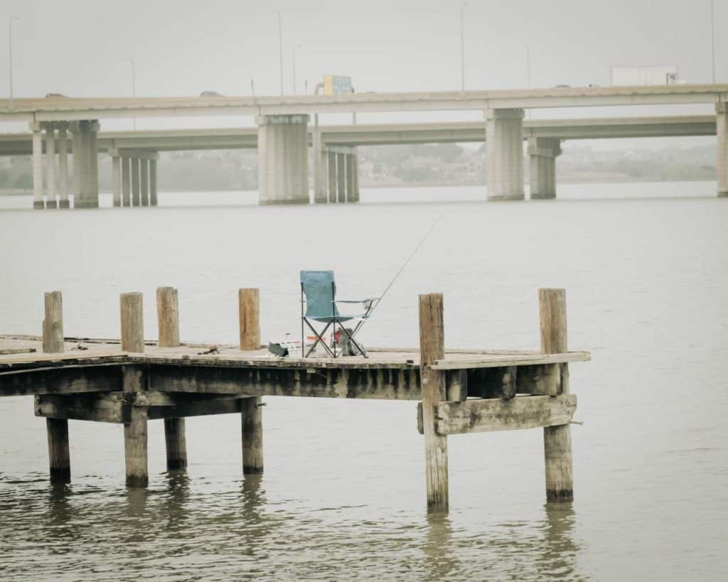 A folding chair and a fishing rod are set up on a wooden pier with a highway in the background