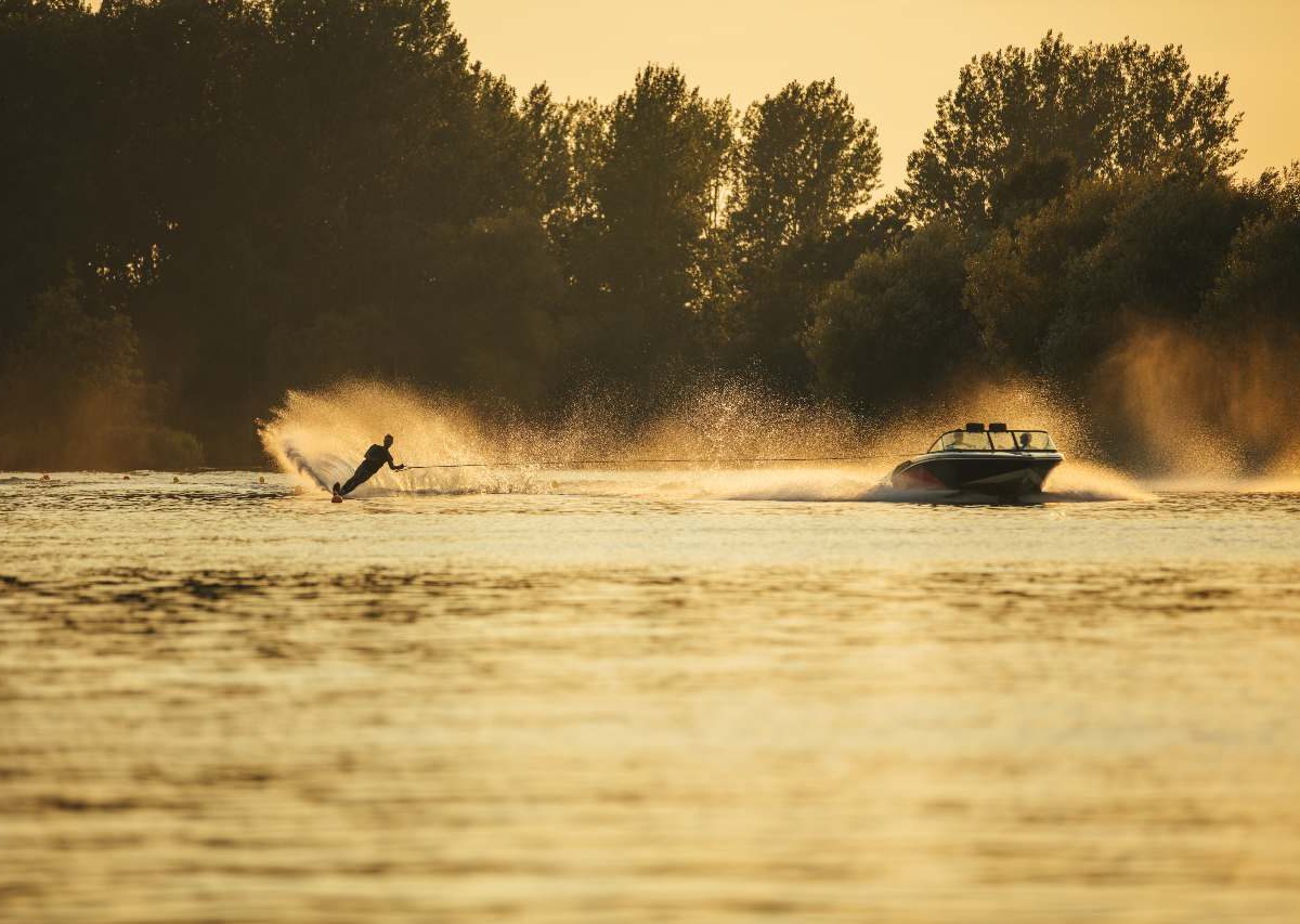 A boat pulls a water skier behind it on a lake with trees in the background, spraying water into the air