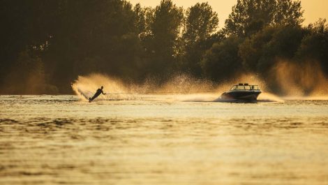 A boat pulls a water skier behind it on a lake with trees in the background, spraying water into the air