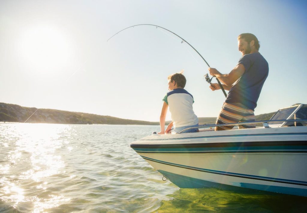 A father and son stand on a boat while the dad fishes in open water.