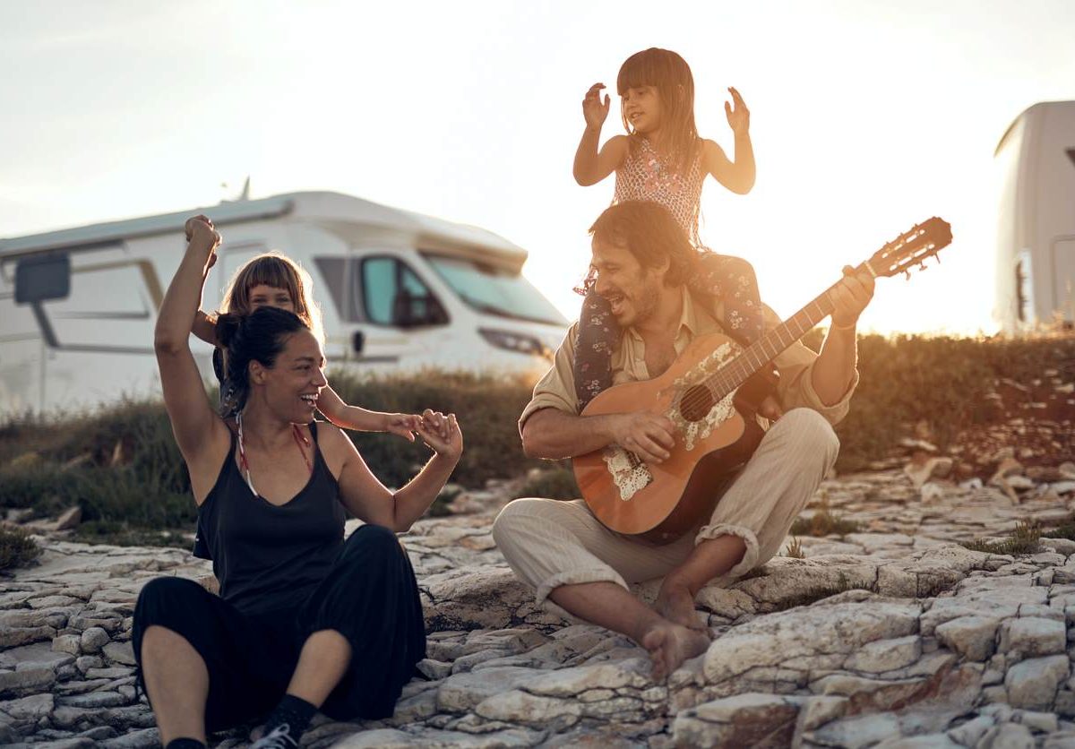 A family of four, traveling in an RV with kids, sits on stones and plays a guitar.