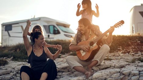 A family of four, traveling in an RV with kids, sits on stones and plays a guitar.