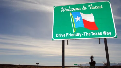 A green road sign that says, “Welcome to Texas. Drive friendly—The Texas Way” marks the Texas state line on a remote country road.