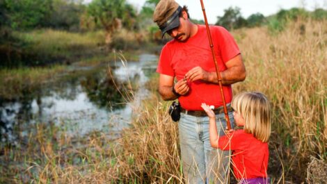 A father baits the hook while the daughter holds the fishing rod beside a pond.