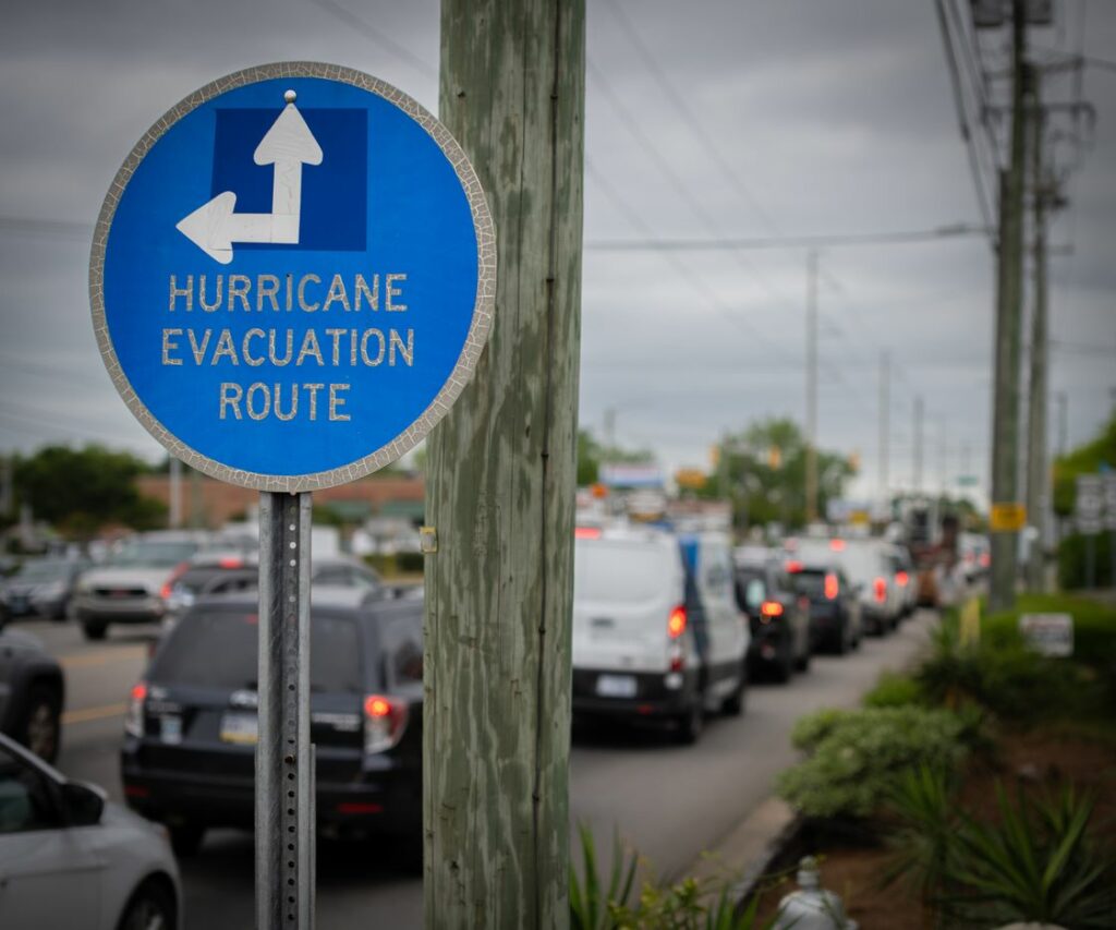 A hurricane evacuation route sign points forward and left as cars wait in traffic.