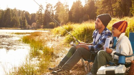 A smiling father and son fish by a river during a fall sunny day.