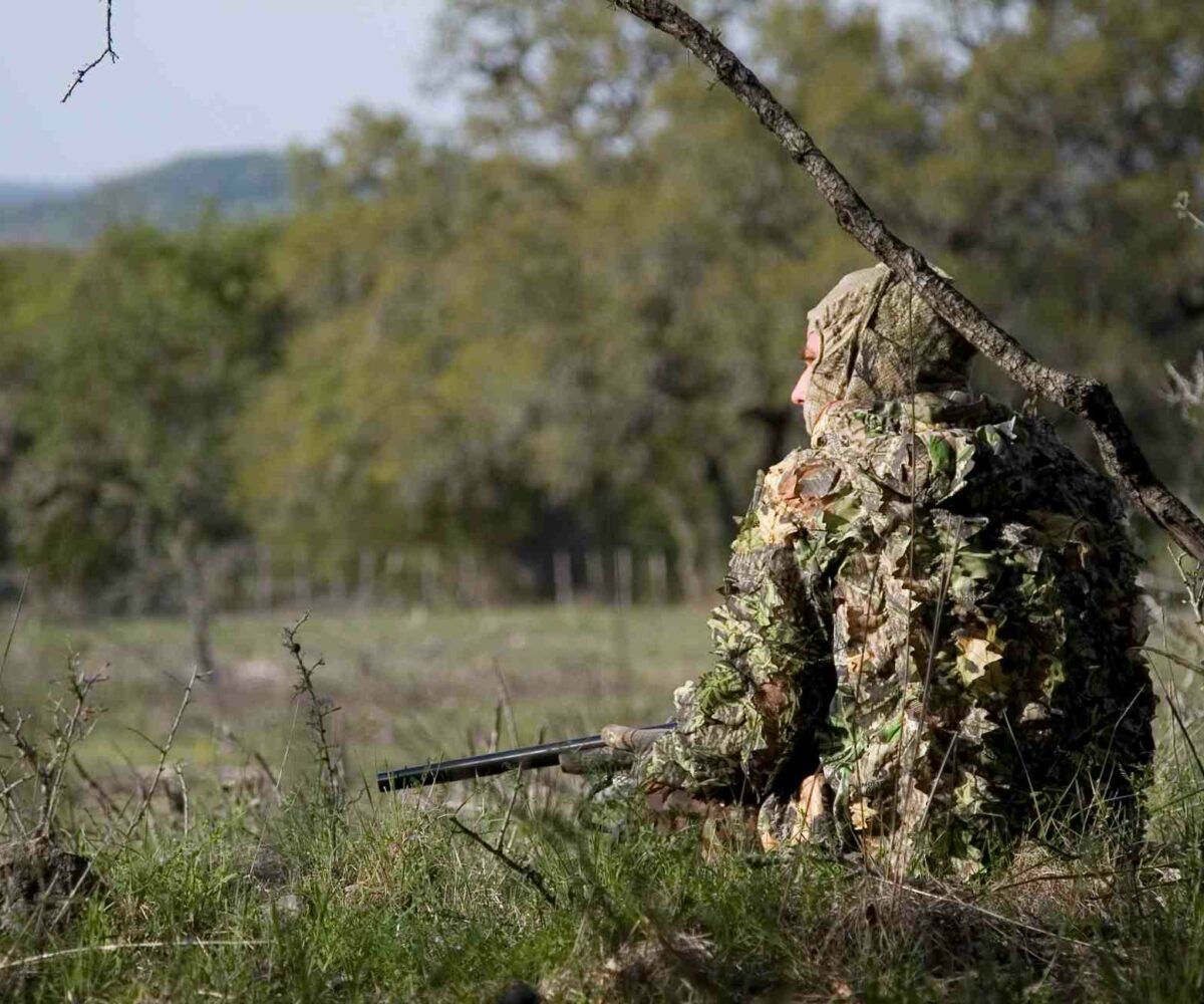 A camouflaged Texas hunter sits on the ground among tall grass.