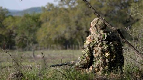 A camouflaged Texas hunter sits on the ground among tall grass.