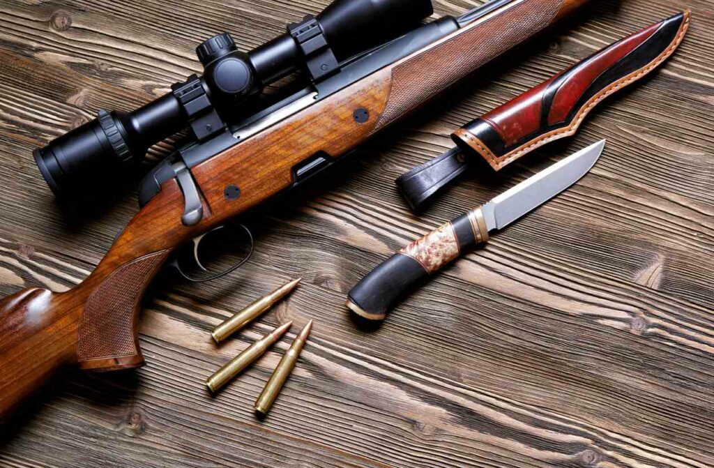 Hunting gear, including a rifle, bullets, and knives, sits on a wooden table.